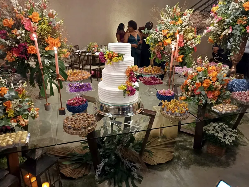 Mesa de vidro com um bolo branco, arranjos de flores laranjas e velas